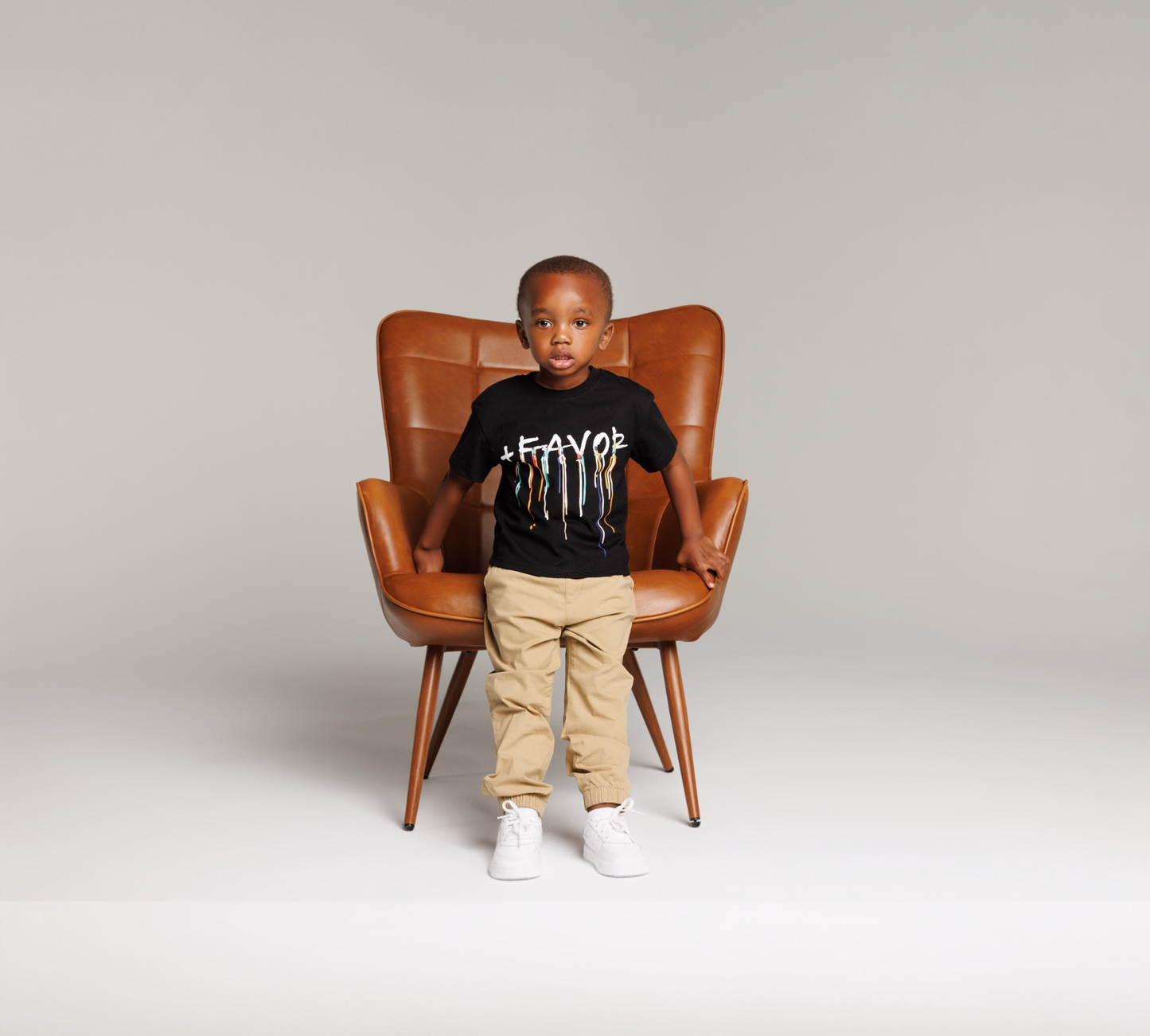Child sitting on a brown leather chair wearing a black t-shirt with white text and beige pants.